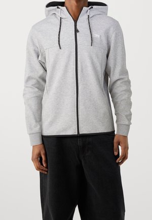 Homme portant un sweat à capuche zippé gris clair avec des cordons de serrage noirs et un pantalon noir, se tenant devant un fond clair uni.