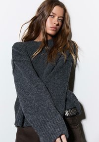 Tmavosivý pletený sveter so širokými rukávmi a lemom siahajúcim po boky. Má textúrovaný povrch a kontrastný čierny opasok so striebornými detailmi.