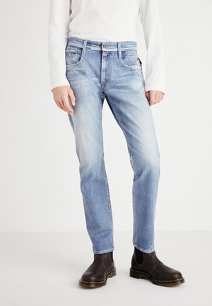 Personne portant un jean slim bleu clair avec des détails délavés et des bottines en cuir marron foncé, debout devant un fond blanc.