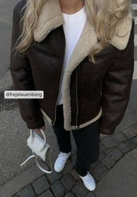 Brun læderjakke med beige shearling-krave, lynlåslukning, sorte bukser, hvide sneakers og lille hvid håndtaske. Teksturerede detaljer synlige.