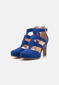 Blue suede high-heeled shoes feature cut-out designs and adjustable ankle straps, with a smooth beige interior and a sturdy wooden heel.