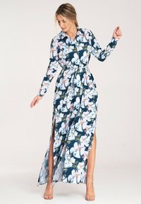 Kwiatowa sukienka maxi z ciemnoniebieskim tłem i białymi kwiatami, długimi rękawami, kołnierzykiem oraz paskiem w talii. Bocznie rozcięcia odsłaniają nogi.