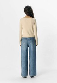 Maglietta a maniche lunghe beige a coste abbinata a jeans a gamba larga blu chiaro con tasche posteriori, vista da dietro su uno sfondo neutro.