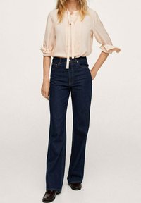 Frau trägt eine hellrosa Bluse mit aufgerollten Ärmeln und dunkelblaue Jeans mit hohem Bund und weitem Bein, steht mit einer Hand auf der Hüfte.