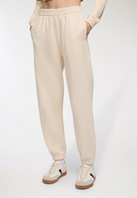 Pantaloni della tuta color crema con vita elasticizzata e orli a costina. Texture morbida con tasche laterali, abbinati a scarpe da ginnastica bianche.