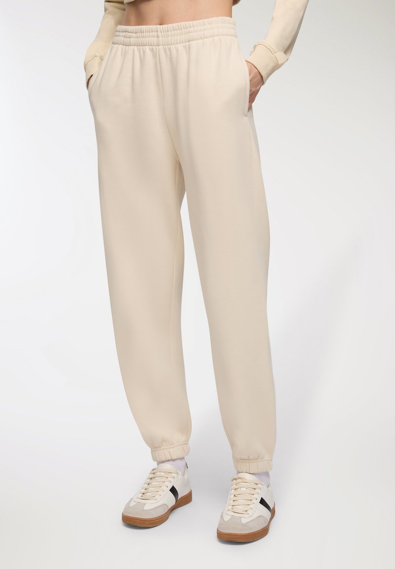 Pantaloni della tuta color crema con vita elasticizzata e orli a costina. Texture morbida con tasche laterali, abbinati a scarpe da ginnastica bianche.