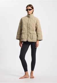 Beige gesteppte Jacke mit hohem Kragen, vorderem Knopfverschluss und zwei großen Taschen. Über schwarzen Leggings getragen, barfuß.