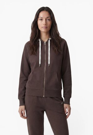 Femme aux longs cheveux foncés portant un sweat à capuche marron zippé avec des cordons blancs et un pantalon marron assorti sur fond blanc.