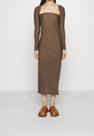 Femme portant une robe midi marron à carreaux ajustée, avec des manches longues et un col carré, associée à des chaussures marron sans lacets.