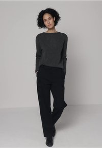 Harmaa neulepaita, jossa pyöreä pääntie, yhdistettynä väljiin mustiin housuihin; malli seisoo neutraalia taustaa vasten.