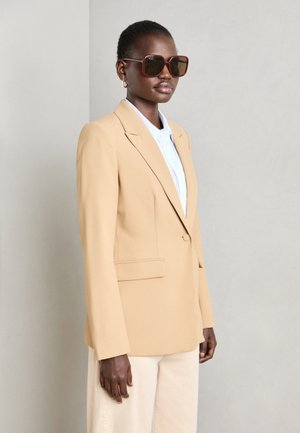 Beige saka na míru s jedním knoflíkem, klopovými límci a bočními kapsami, nošené přes světle modrou košili. Hnědé oversized sluneční brýle dotvářejí vzhled.