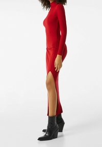 Röd långärmad klänning med hög krage, med sidosprund och en figurnära design. Accenterad med svarta ankelstövlar med klack.