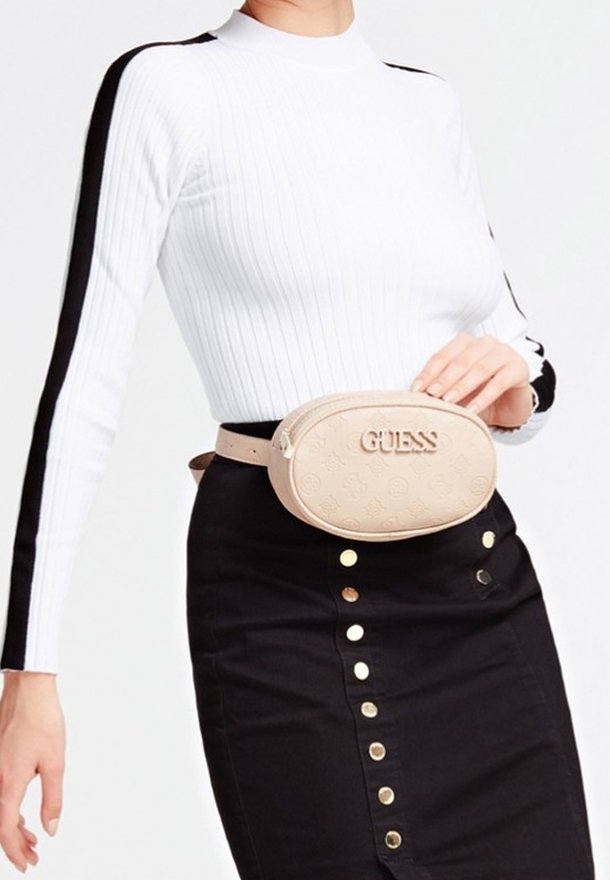 Femme portant un pull blanc côtelé avec des rayures noires et une jupe noire boutonnée, accessoirisée d'un sac ceinture beige Guess.