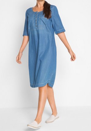 Donna che indossa un vestito in denim blu fino al ginocchio con maniche a tre quarti e bottoni sul davanti, camminando con scarpe bianche slip-on.