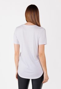 Vrouw met lang, steil bruin haar, gekleed in een eenvoudige witte shirt met korte mouwen en zwarte broek, gezien vanaf de achterkant.