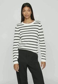 Suéter listrado em branco e preto, com um decote redondo, mangas longas e um design de malha texturizada. Combinado com calças pretas.