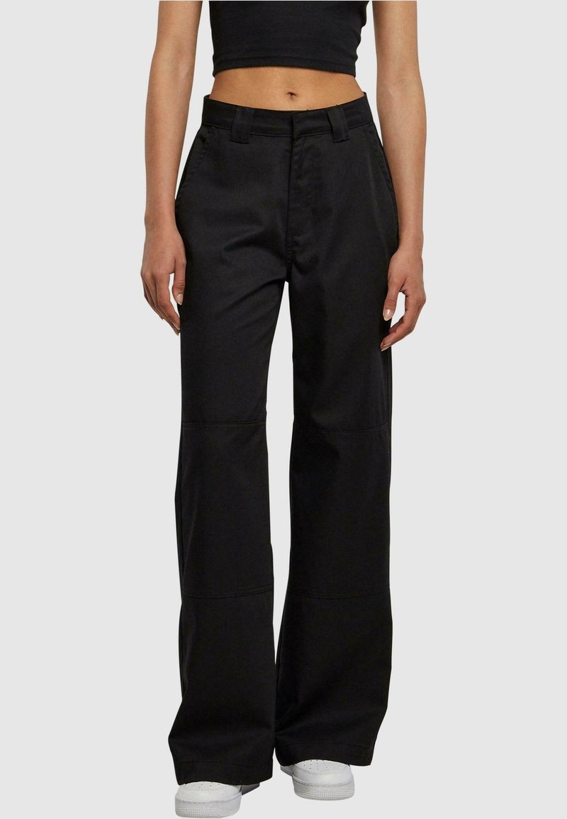 Personne portant un pantalon noir taille haute à jambes larges avec des détails de couture, des baskets blanches et un top court noir sur un fond uni.