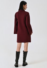 Abito lavorato a maglia bordeaux con colletto alto e maniche a campana, abbinato a stivali neri in pelle al ginocchio con dettagli dorati.