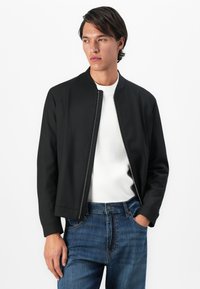 Giovane uomo con una giacca nera con zip sopra una camicia bianca e jeans blu, in piedi con una mano in tasca, che guarda di lato.