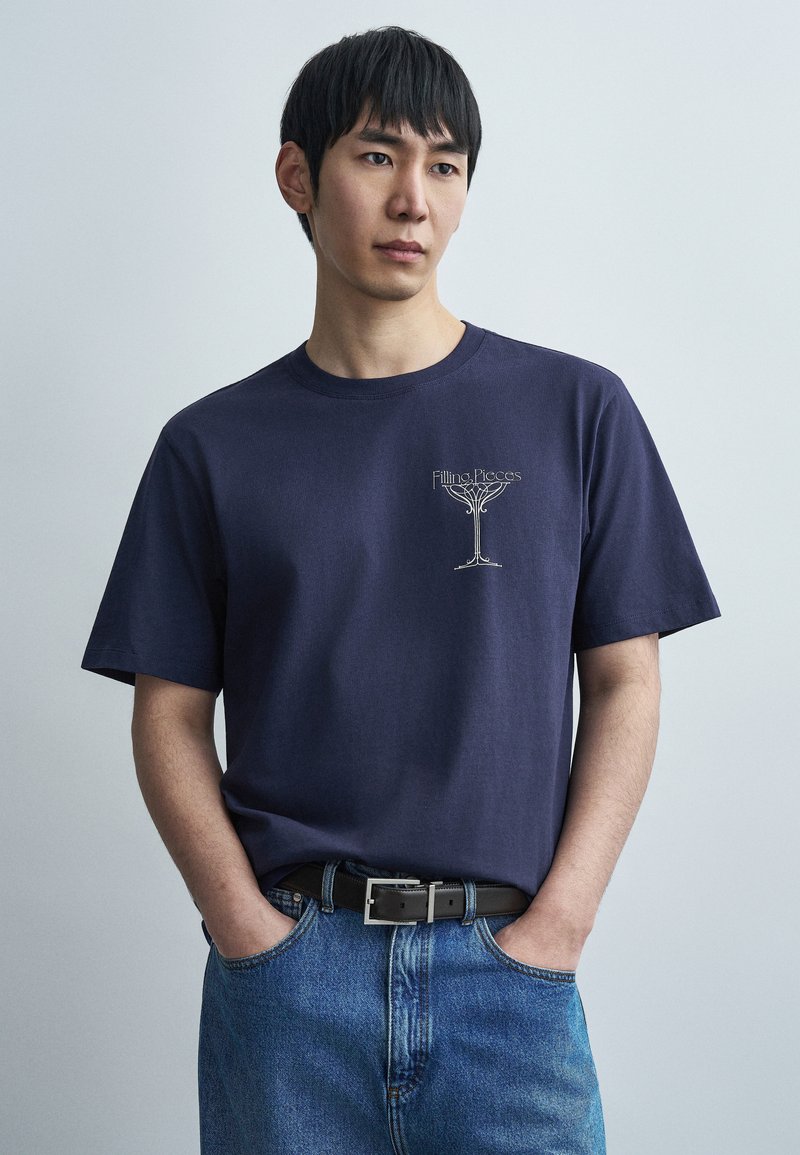 Μπλε βαμβακερό t-shirt με στρογγυλή λαιμόκοψη και κοντά μανίκια, με ανοιχτόχρωμο σχέδιο λογότυπου στο στήθος· συνδυασμένο με μπλε τζιν παντελόνι.