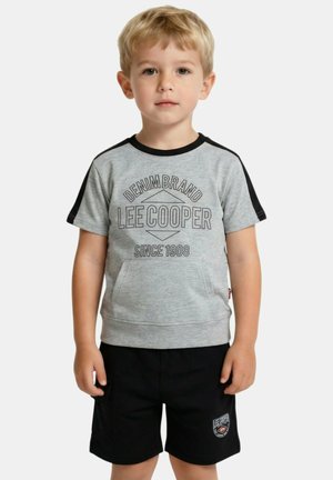 Jeune garçon blond debout, portant un t-shirt gris Lee Cooper avec des manches noires et un short noir avec un patch logo Lee Cooper.
