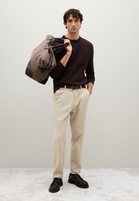 Maglione marrone strutturato, pantaloni beige, scarpe nere. Porta una borsa marrone con motivo e dettagli neri. Sfondo neutro.