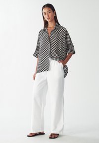 Košile s černobílým vzorem, krátkými rukávy a volným střihem, spárovaná s bílými kalhotami s širokými nohavicemi a černými plochými sandály.