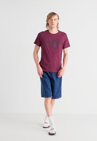 Bordeauxrotes T-Shirt mit schwarzem Levi's-Logo, kombiniert mit knielangen Denim-Shorts, weißen Socken und schwarzen Sandalen. Das Model steht vor einem schlichten Hintergrund.