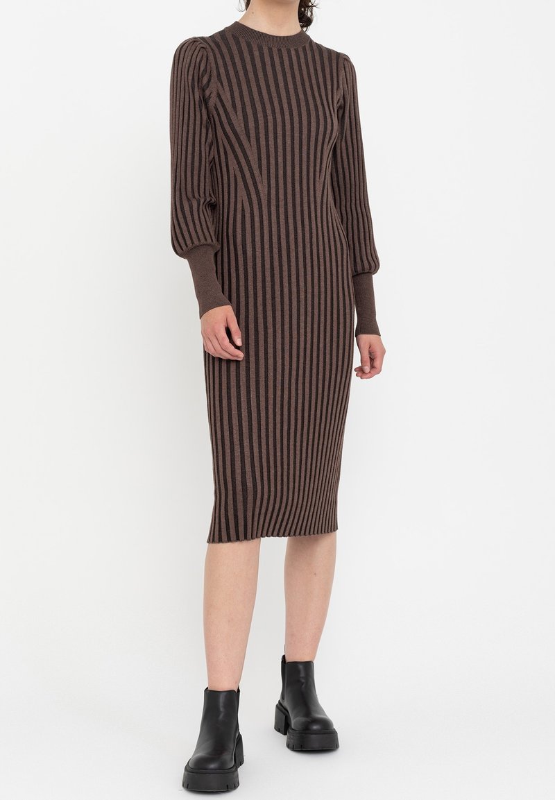 Femme portant une robe midi rayée verticale marron et noire avec des manches longues bouffantes et des bottines noires.
