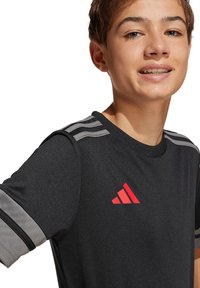 T-shirt de sport Adidas noir avec des bandes grises sur les épaules. Présente un logo rouge sur la poitrine. Texture lisse, manches courtes, design col rond.