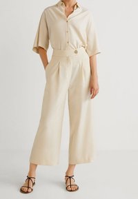Beige button-up overhemd met korte mouwen, gecombineerd met wijd-uitlopende beige broek met plooien en een knoop aan de voorkant. Zwarte sandalen met bandjes.