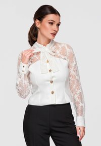 Femme en blouse blanche en dentelle avec nœud papillon et boutons dorés, associée à un pantalon noir taille haute, posant devant un fond uni.