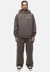 Set di felpa con cappuccio e pantaloni in cotone marrone scuro, con tasca frontale e dettagli del logo in bianco. Vestibilità rilassata con orli a coste.