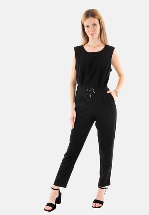 Sort jumpsuit med rund halsudskæring, ærmeløs design, snoretræk i taljen og tapered ben. Modellen bærer sorte hæle sandaler.