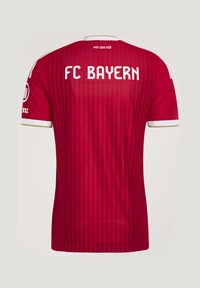 Κόκκινη φανέλα ποδοσφαίρου FC Bayern με λευκό κείμενο "FC BAYERN" στην πλάτη και "MIA SAN MIA" κοντά στη λαιμόκοψη, με διακριτικές κάθετες ρίγες.