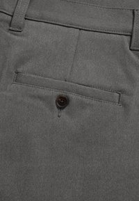 Gros plan sur un pantalon en tissu gris montrant une poche arrière passepoilée fermée par un seul bouton foncé.