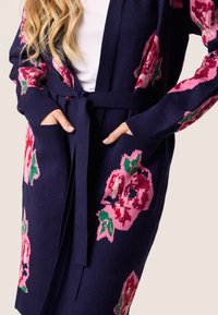 Marine gebreide cardigan met roze bloemenpatronen, een gestrikt middel, een open voorkant en zijzakken, voorzien van geribbelde manchetten voor extra textuur.