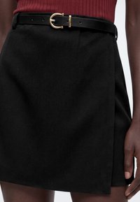 Jupe noire avec une ceinture noire à boucle dorée, portée avec un haut rouge côtelé, montrée de la taille jusqu'au milieu de la cuisse avec une main détendue sur le côté.