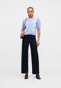 Γυναίκα με πλεγμένα μαλλιά, που φοράει γαλάζιο πουλόβερ με κοντά μανίκια και διαμάντι σχέδια, σκούρο παντελόνι φαρδύ, μαύρα παπούτσια με τακούνι και τσάντα ώμου.