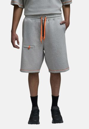 Homme portant un short gris jusqu'aux genoux avec des cordons et des bordures orange, des chaussettes noires et des baskets noires, debout avec une main dans la poche.