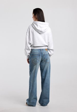 Vrouw met lang donker haar, gekleed in een witte hoodie en losse blauwe spijkerbroek, staand met haar rug naar de camera tegen een effen achtergrond.