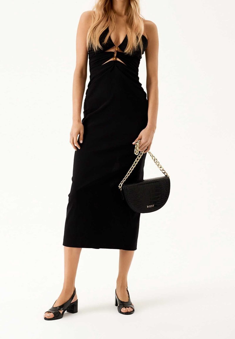 Femme en robe midi noire sans manches avec découpes, tenant un sac à main noir avec une bandoulière chaîne dorée, portant des sandales noires à talons.