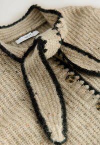 Beige Strickcardigan mit strukturiertem Muster, schwarzem Besatz und einer Schleifenverschluss am Hals. Verfügt über runde Knöpfe an der Vorderseite.