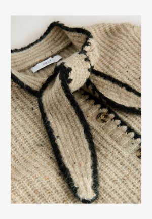 Cárdigan de punto beige con un patrón texturizado, ribete negro y un lazo en el cuello. Presenta botones redondos a lo largo del frente.