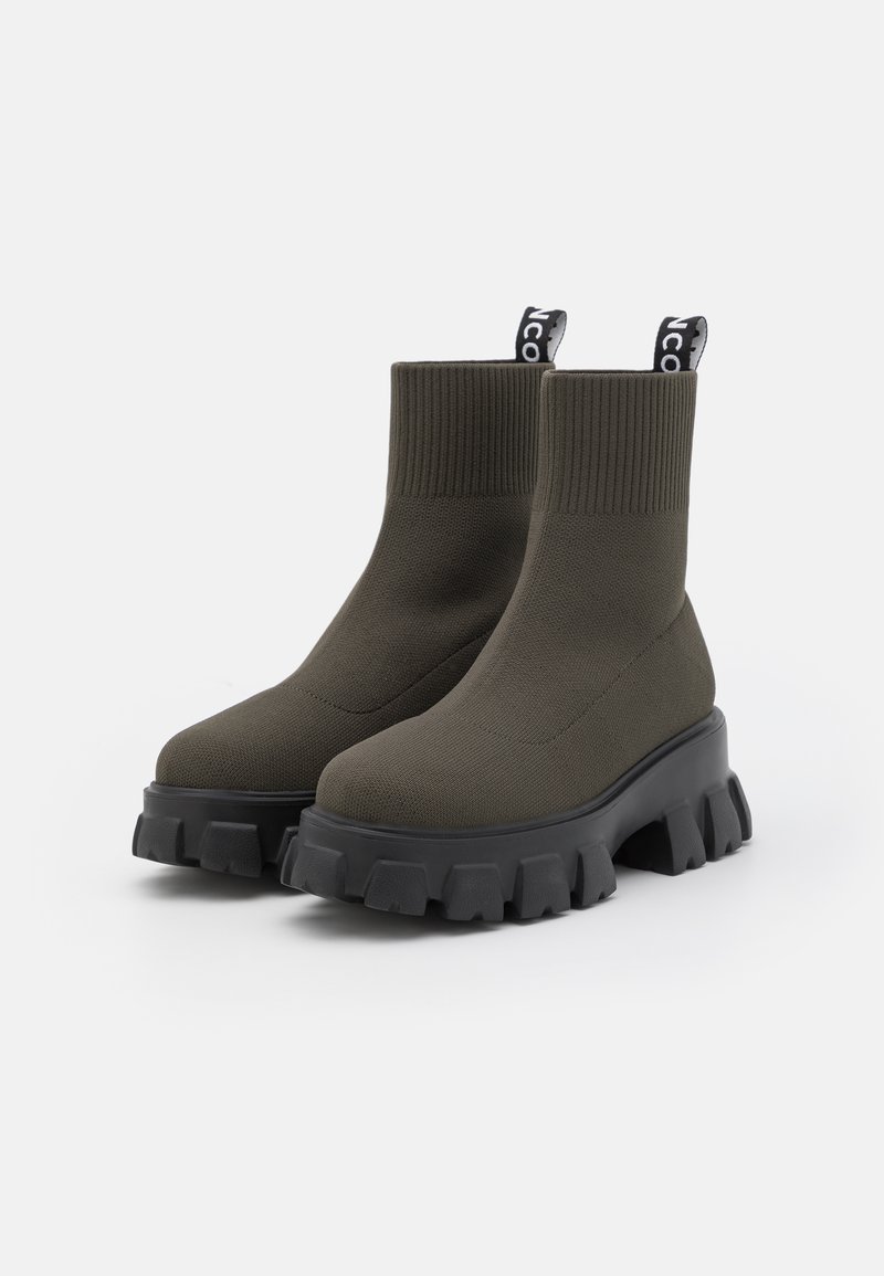 Bottes de cheville vert olive foncé avec un dessus en tricot côtelé, semelle extérieure en caoutchouc noir chunky, et tirant avec logo blanc.