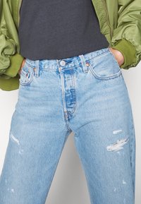 Jeans de denim azul claro com um corte relaxado, apresentando detalhes desgastados e uma cintura alta. Combinados com uma camisa cinza escura e uma jaqueta bomber verde.