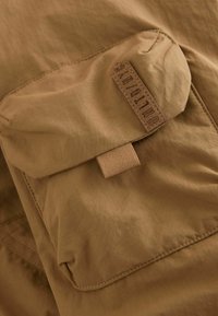Tessuto khaki con una superficie liscia presenta una tasca superiore a patta e un'etichetta con testo in rilievo. Design semplice, senza motivi o accenti visibili.