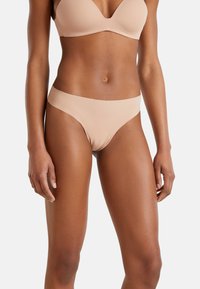 Vrouw in naadloze huidkleurige beha en bijpassende bikinislip, staande met ontspannen armen langs haar lichaam op een witte achtergrond.