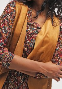 Béžová faux semišová vesta s cvočky přehozená přes květovanou halenku s odstíny červené, modré a žluté. Viditelné jsou dlouhé rukávy.