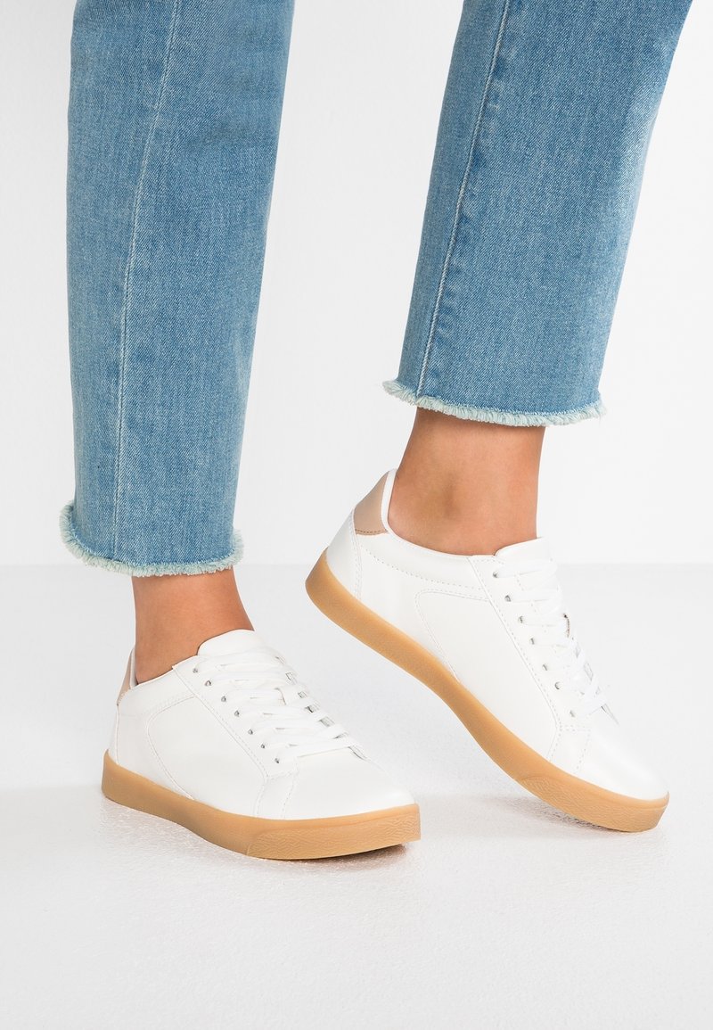 Jean bleu clair longueur cheville avec ourlets effilochés, porté avec des baskets basses blanches avec des accents beiges au talon et des semelles en caoutchouc gomme.